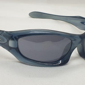 May include: Blue-tinted sunglasses with dark gray lenses. The sunglasses have a curved frame and arms, with a silver emblem on the side. The lenses are a dark gray color, and the overall style is sporty.
