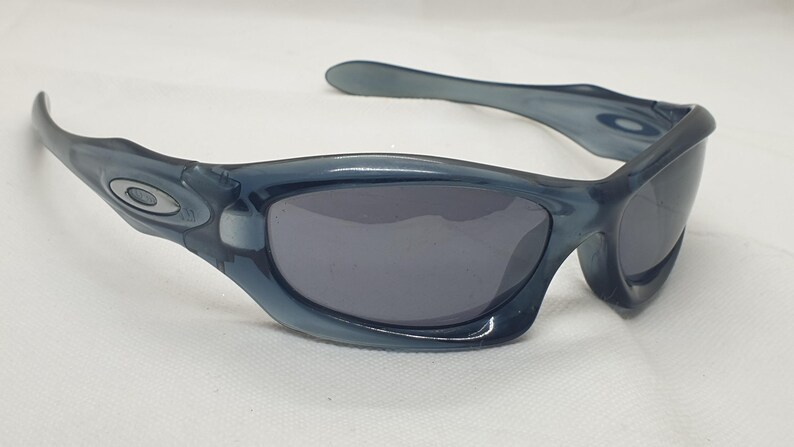 May include: Blue-tinted sunglasses with dark gray lenses. The sunglasses have a curved frame and arms, with a silver emblem on the side. The lenses are a dark gray color, and the overall style is sporty.