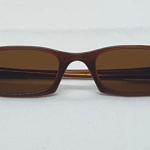 Pode incluir: &Oacute;culos de sol castanhos com arma&ccedil;&atilde;o retangular e lentes tingidas de castanho. As hastes s&atilde;o de um castanho mais escuro com um detalhe azul marinho perto das dobradi&ccedil;as. Os &oacute;culos de sol est&atilde;o sobre um fundo branco.
