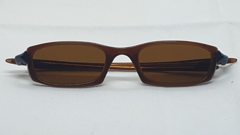 Pode incluir: &Oacute;culos de sol castanhos com arma&ccedil;&atilde;o retangular e lentes tingidas de castanho. As hastes s&atilde;o de um castanho mais escuro com um detalhe azul marinho perto das dobradi&ccedil;as. Os &oacute;culos de sol est&atilde;o sobre um fundo branco.