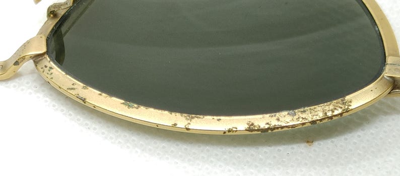 May include: Close-up of vintage sunglasses with a gold-colored metal frame and dark green lenses. The frame shows signs of wear and age, with some discoloration and spots. The lenses are slightly curved and reflect the light.