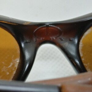 Puede incluir: Primer plano de gafas de sol con montura marr&oacute;n y lentes &aacute;mbar. El puente de las gafas tiene un peque&ntilde;o detalle ovalado. Las lentes muestran algo de desgaste.