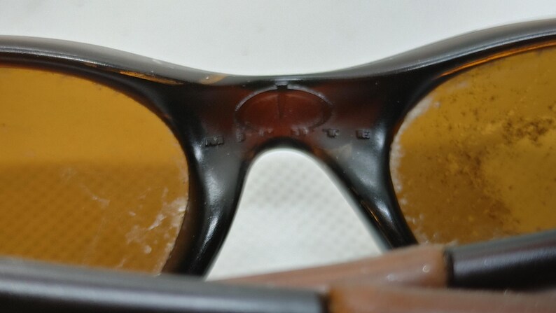 Puede incluir: Primer plano de gafas de sol con montura marr&oacute;n y lentes &aacute;mbar. El puente de las gafas tiene un peque&ntilde;o detalle ovalado. Las lentes muestran algo de desgaste.