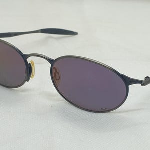 May include: Oval-shaped sunglasses with a dark gray metal frame and purple-tinted lenses. The lenses reflect a gradient of colors. The arms of the sunglasses are also dark gray.