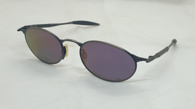 May include: Oval-shaped sunglasses with a dark gray metal frame and purple-tinted lenses. The lenses reflect a gradient of colors. The arms of the sunglasses are also dark gray.