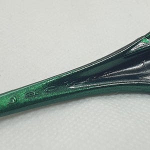 May include: Close-up of a green and black sunglasses arm. The arm is a metallic green with the brand name etched in the side. The lens is visible in the background.