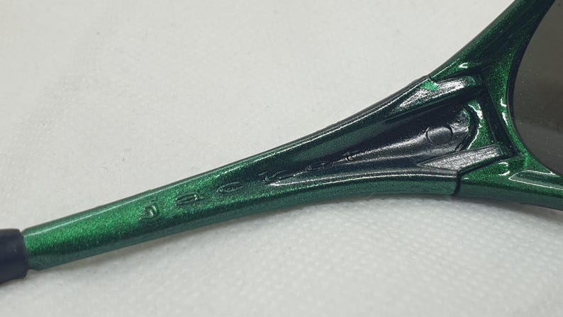 May include: Close-up of a green and black sunglasses arm. The arm is a metallic green with the brand name etched in the side. The lens is visible in the background.