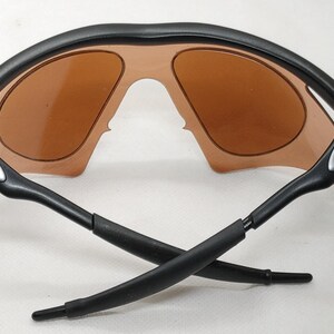 May include: Black sunglasses with brown lenses. The sunglasses have a curved black frame and black arms. The lenses are a light brown colour. The arms cross each other.