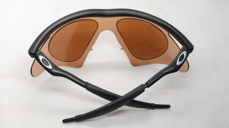 May include: Black sunglasses with brown lenses. The sunglasses have a curved black frame and black arms. The lenses are a light brown colour. The arms cross each other.