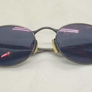 May include: Oval-shaped sunglasses with a dark grey metal frame and dark blue lenses. The sunglasses have a classic design with a bridge and temples. The lenses reflect a reddish hue.