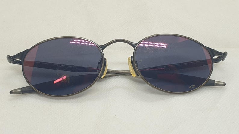May include: Oval-shaped sunglasses with a dark grey metal frame and dark blue lenses. The sunglasses have a classic design with a bridge and temples. The lenses reflect a reddish hue.