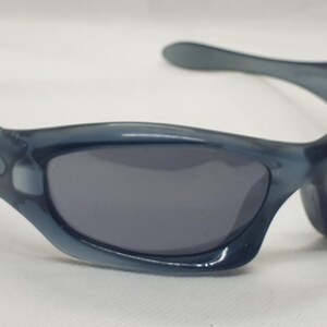 May include: Blue-tinted sunglasses with dark gray lenses. The sunglasses have a curved frame and arms, with the Oakley logo visible on the temples. The lenses are a dark gray color, and the frame is a translucent blue.