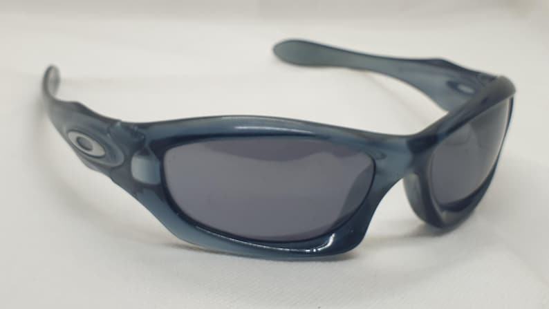 May include: Blue-tinted sunglasses with dark gray lenses. The sunglasses have a curved frame and arms, with the Oakley logo visible on the temples. The lenses are a dark gray color, and the frame is a translucent blue.
