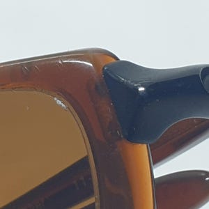 Pode incluir: Close-up de &oacute;culos de sol. A arma&ccedil;&atilde;o &eacute; marrom quente, com uma dobradi&ccedil;a azul marinho escuro. As lentes s&atilde;o de cor marrom claro. Os &oacute;culos de sol est&atilde;o em um fundo branco.