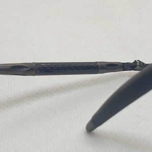 May include: Close-up of a pair of sunglasses. The frame is dark gray with a black carbon fiber-like section at the hinge. The lens is a dark blue color. The arm of the glasses extends to the right.