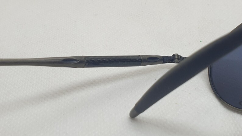 May include: Close-up of a pair of sunglasses. The frame is dark gray with a black carbon fiber-like section at the hinge. The lens is a dark blue color. The arm of the glasses extends to the right.