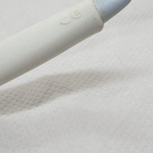 K&ouml;nnte beinhalten: Nahaufnahme eines wei&szlig;en Kosmetikstifts mit hellblauer Spitze. Der Stift hat eine glatte, matte Oberfl&auml;che und liegt auf einer wei&szlig;en, strukturierten Oberfl&auml;che. Die Buchstaben "CE" sind auf den Stift gedruckt.