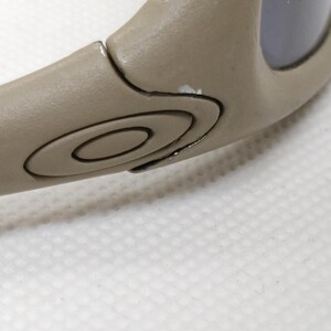 Puede incluir: Primer plano de una patilla de gafas de sol de color beige. El brazo presenta un dise&ntilde;o circular con el logotipo de la marca y algunas marcas de pintura blanca. La lente es de color oscuro. Las gafas de sol est&aacute;n sobre una superficie blanca.