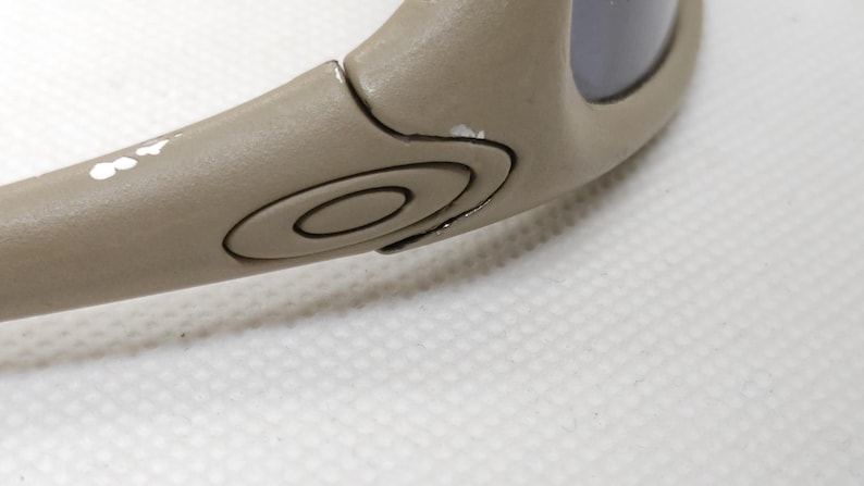 Puede incluir: Primer plano de una patilla de gafas de sol de color beige. El brazo presenta un dise&ntilde;o circular con el logotipo de la marca y algunas marcas de pintura blanca. La lente es de color oscuro. Las gafas de sol est&aacute;n sobre una superficie blanca.