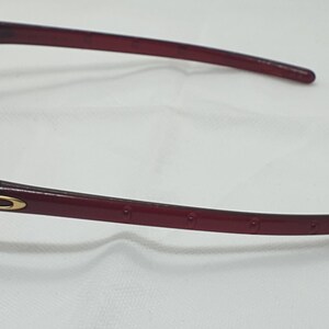 May include: Maroon sunglasses with a gold accent on the temple. The lenses are light brown. The frame has a curved design and is made of a glossy material. The arms have small, evenly spaced holes.