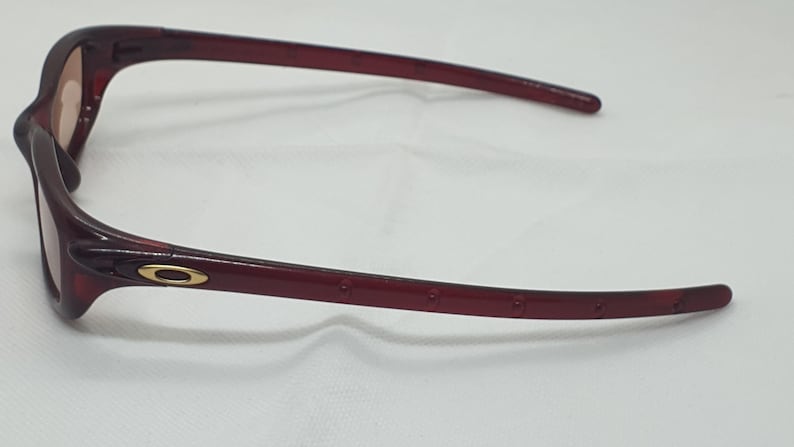 May include: Maroon sunglasses with a gold accent on the temple. The lenses are light brown. The frame has a curved design and is made of a glossy material. The arms have small, evenly spaced holes.