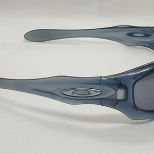 May include: Blue-tinted sunglasses with a wraparound design. The sunglasses have a dark gray tint and a silver logo on the frame. The arms curve for a secure fit. These are a sporty accessory.