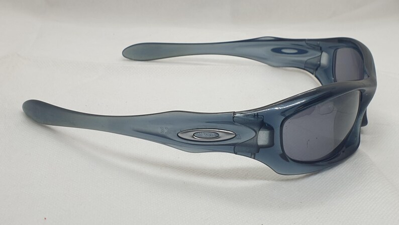 May include: Blue-tinted sunglasses with a wraparound design. The sunglasses have a dark gray tint and a silver logo on the frame. The arms curve for a secure fit. These are a sporty accessory.