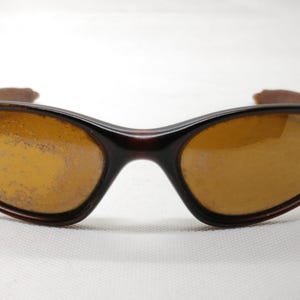 Puede incluir: Gafas de sol marrones con lentes &aacute;mbar. Las gafas tienen una montura marr&oacute;n oscuro y un dise&ntilde;o curvado. Las lentes parecen estar ligeramente rayadas. Las patillas de las gafas son de color marr&oacute;n claro.