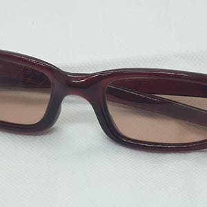 May include: Burgundy sunglasses with a rectangular frame and tinted lenses. The sunglasses have a curved design at the temples. The lenses are a light brown colour. The sunglasses are set against a white background.