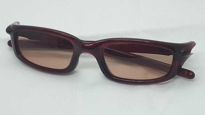 May include: Burgundy sunglasses with a rectangular frame and tinted lenses. The sunglasses have a curved design at the temples. The lenses are a light brown colour. The sunglasses are set against a white background.