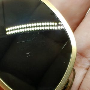 May include: Sunglasses with a gold-colored frame and dark lenses. The sunglasses have a teardrop shape and are held in a hand. The lenses have some scratches.