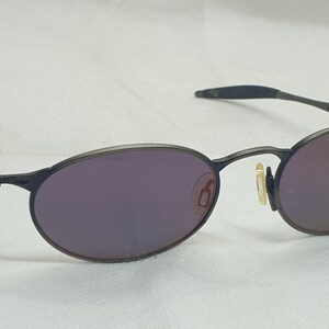 May include: Oval-shaped sunglasses with a dark gray metal frame and purple-tinted lenses. The lenses have a reflective quality. The arms of the glasses are thin and extend to the ear pieces.