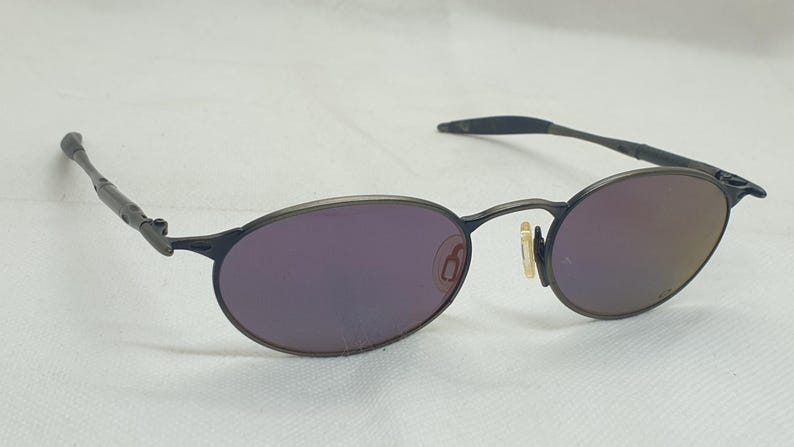 May include: Oval-shaped sunglasses with a dark gray metal frame and purple-tinted lenses. The lenses have a reflective quality. The arms of the glasses are thin and extend to the ear pieces.