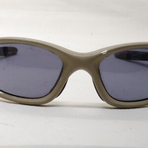 Puede incluir: Gafas de sol color taupe con lentes oscuras. Las gafas tienen una montura curvada y un peque&ntilde;o detalle decorativo en el lateral. Las lentes son de color gris oscuro.