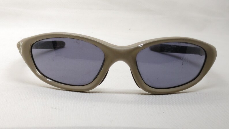 Puede incluir: Gafas de sol color taupe con lentes oscuras. Las gafas tienen una montura curvada y un peque&ntilde;o detalle decorativo en el lateral. Las lentes son de color gris oscuro.