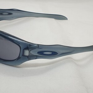 May include: A pair of blue-grey sunglasses with a curved frame and dark lenses. The sunglasses have a sporty design and the Oakley logo is visible on the temples. The glasses are set against a white background.