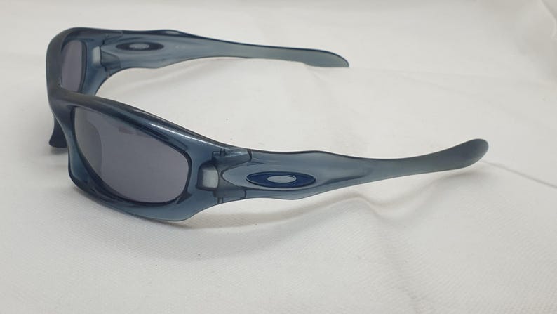 May include: A pair of blue-grey sunglasses with a curved frame and dark lenses. The sunglasses have a sporty design and the Oakley logo is visible on the temples. The glasses are set against a white background.