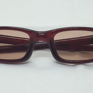 May include: Burgundy sunglasses with a rectangular frame and tinted lenses. The glasses are folded on a white surface. The text "MADE IN U.S.A." is visible on the frame. A stylish accessory for eye protection.