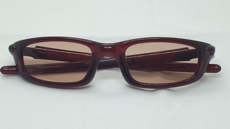 May include: Burgundy sunglasses with a rectangular frame and tinted lenses. The glasses are folded on a white surface. The text "MADE IN U.S.A." is visible on the frame. A stylish accessory for eye protection.