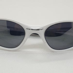 Puede incluir: Gafas de sol blancas con lentes oscuras. Las gafas tienen una forma ovalada elegante y una montura blanca. El logotipo de Oakley es visible en el puente. Las gafas est&aacute;n dise&ntilde;adas para proteger los ojos del sol.