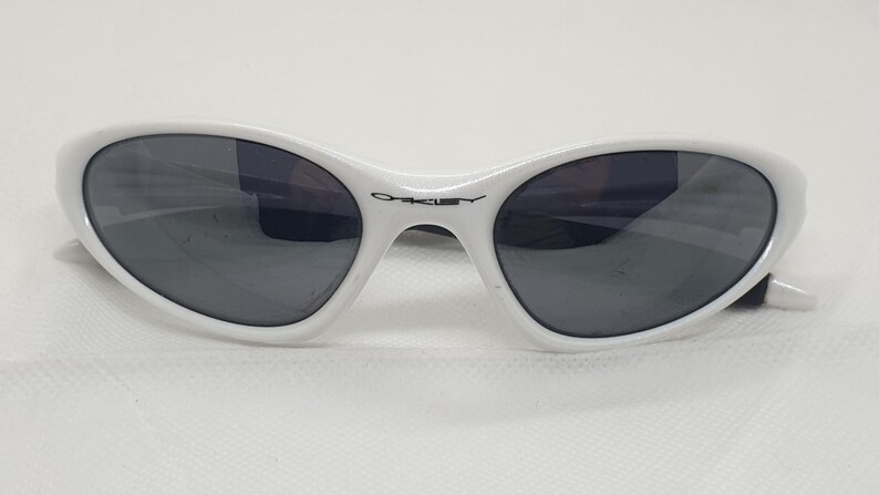 Puede incluir: Gafas de sol blancas con lentes oscuras. Las gafas tienen una forma ovalada elegante y una montura blanca. El logotipo de Oakley es visible en el puente. Las gafas est&aacute;n dise&ntilde;adas para proteger los ojos del sol.