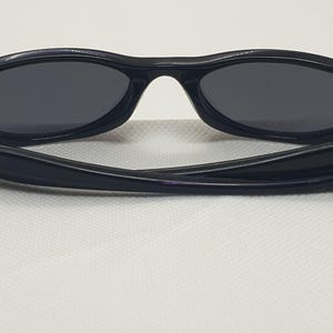 May include: Black sunglasses with oval lenses and a curved frame. The temples have silver accents. The sunglasses are set against a white background.