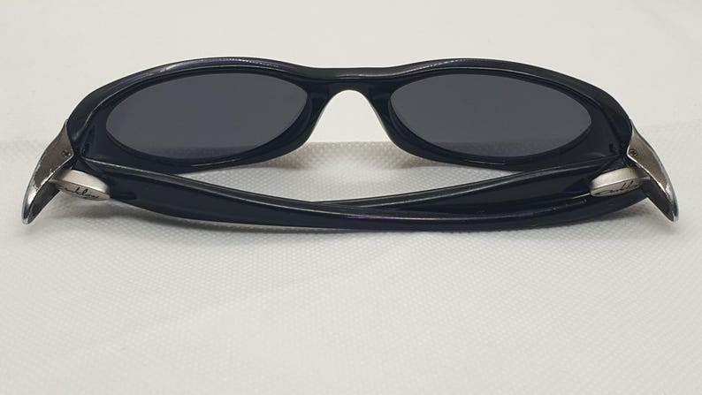 May include: Black sunglasses with oval lenses and a curved frame. The temples have silver accents. The sunglasses are set against a white background.