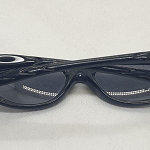 Puede incluir: Gafas de sol negras con un dise&ntilde;o elegante y moderno. Las gafas de sol tienen un acabado brillante y una forma de montura distintiva. Las lentes son oscuras y las patillas tienen un acento met&aacute;lico. Las gafas se muestran sobre un fondo blanco.
