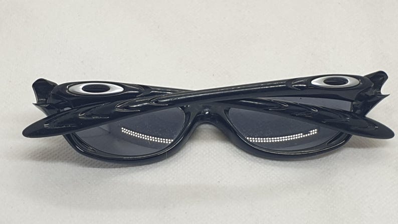 Puede incluir: Gafas de sol negras con un dise&ntilde;o elegante y moderno. Las gafas de sol tienen un acabado brillante y una forma de montura distintiva. Las lentes son oscuras y las patillas tienen un acento met&aacute;lico. Las gafas se muestran sobre un fondo blanco.