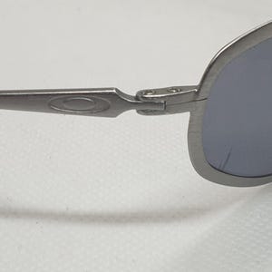 K&ouml;nnte beinhalten: Nahaufnahme einer silberfarbenen Sonnenbrille mit dunkelgrauen Gl&auml;sern. Die Sonnenbrille hat ein schlankes, modernes Design mit einem gebogenen Rahmen und schwarzen B&uuml;gelenden. Das Oakley-Logo ist auf dem B&uuml;gel sichtbar.