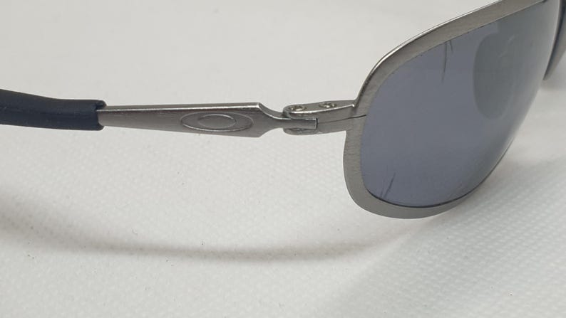 K&ouml;nnte beinhalten: Nahaufnahme einer silberfarbenen Sonnenbrille mit dunkelgrauen Gl&auml;sern. Die Sonnenbrille hat ein schlankes, modernes Design mit einem gebogenen Rahmen und schwarzen B&uuml;gelenden. Das Oakley-Logo ist auf dem B&uuml;gel sichtbar.