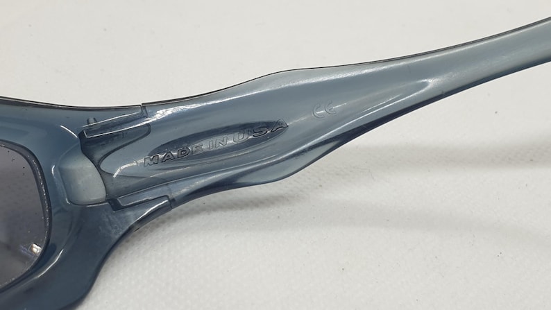 May include: Close-up of a pair of gray sunglasses. The sunglasses have a sleek, modern design with a curved frame. The text "MADE IN USA" is visible on the arm of the sunglasses.