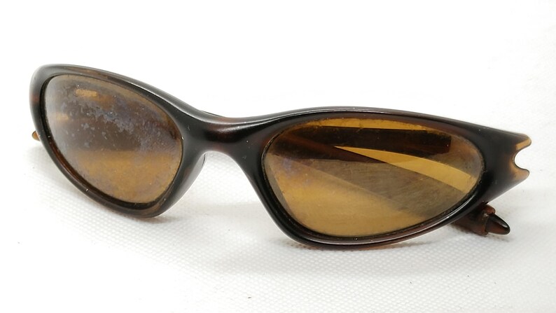 Puede incluir: Gafas de sol marrones con estampado de carey. Las lentes son de color &aacute;mbar c&aacute;lido y las monturas tienen un dise&ntilde;o elegante y curvado. Sobre un fondo blanco.