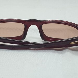 May include: Maroon sunglasses with a rectangular frame and tinted lenses. The arms are folded, revealing a gold-coloured logo on the hinge. The glasses are set against a white background.
